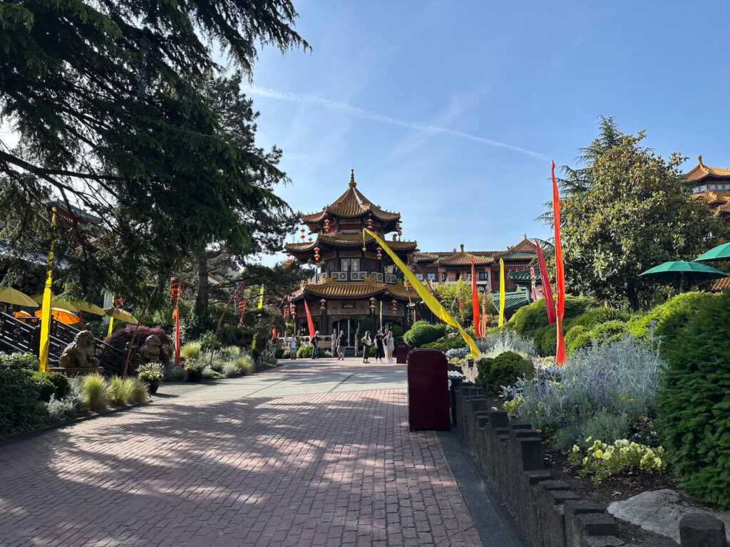 Ein gepflasterter Weg führt durch die China Town des Phantasialands, vorbei an Blumenbeeten und bunten Fahnen zu einem chinesischen Pagoden-Gebäude.