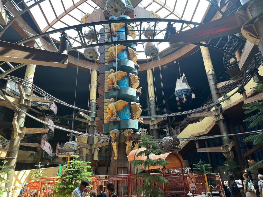 
Blick in die große Kuppelhalle der Attraktion Winja's mit einer hohen, kunstvoll verzierten Mittelsäule voller Enten-Figuren und verschlungenen Schienen unter einem Glasdach.