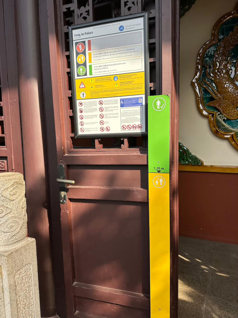 Ein grün-gelber Messpfosten mit 1,20-m-Markierung steht neben einer Infotafel mit den Nutzungsbedingungen der Attraktion Feng Ju Palace.
