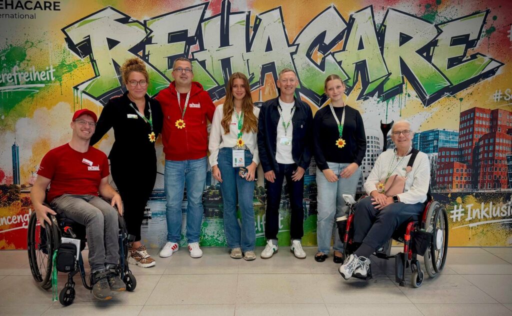Schild Rehacare und viele Menschen davor für ein Gruppenfoto. Man sieht T-Shirts mit dem Logo von Freizeitpark Erlebnis