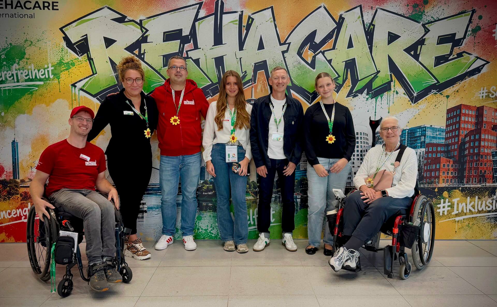 Schild Rehacare und viele Menschen davor für ein Gruppenfoto. Man sieht T-Shirts mit dem Logo von Freizeitpark Erlebnis